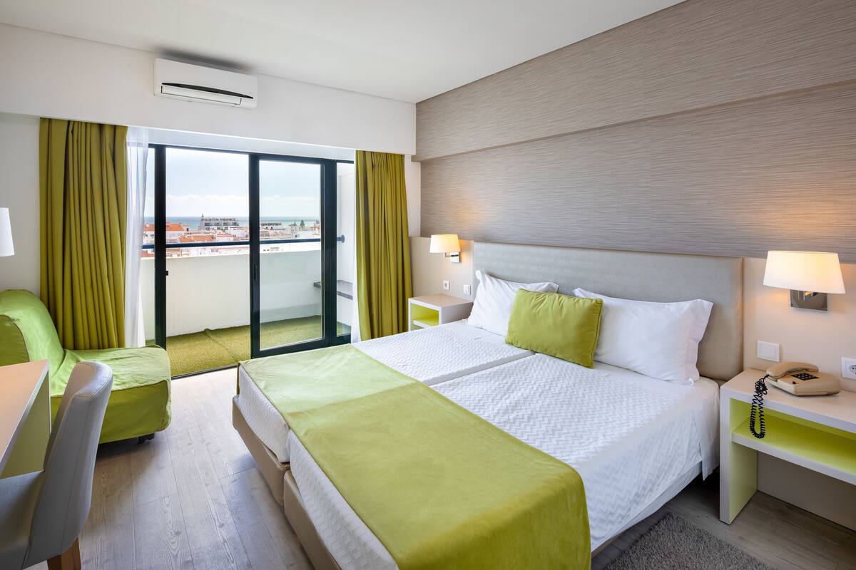 A hotel room with a large bed, a desk, a chair, and a view of the city through a glass door.