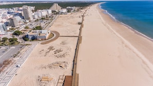 An aerial view of a coastal area with a sandy beach, a promenade, and a row of buildings along the shore. - Hotel Navegadores (Vila Real Santo Antonio)