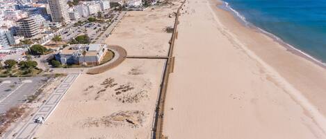 An aerial view of a coastal area with a sandy beach, a promenade, and a row of buildings along the shore.