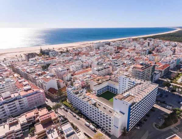 An aerial view of a coastal city with dense urban development, a prominent white building with a blue accent, and a beach in the background. - Hotel Navegadores (Vila Real Santo Antonio)
