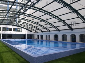 An indoor swimming pool with a glass ceiling and a tiled edge, surrounded by a white building with arched windows. - Hotel Navegadores (Vila Real Santo Antonio)