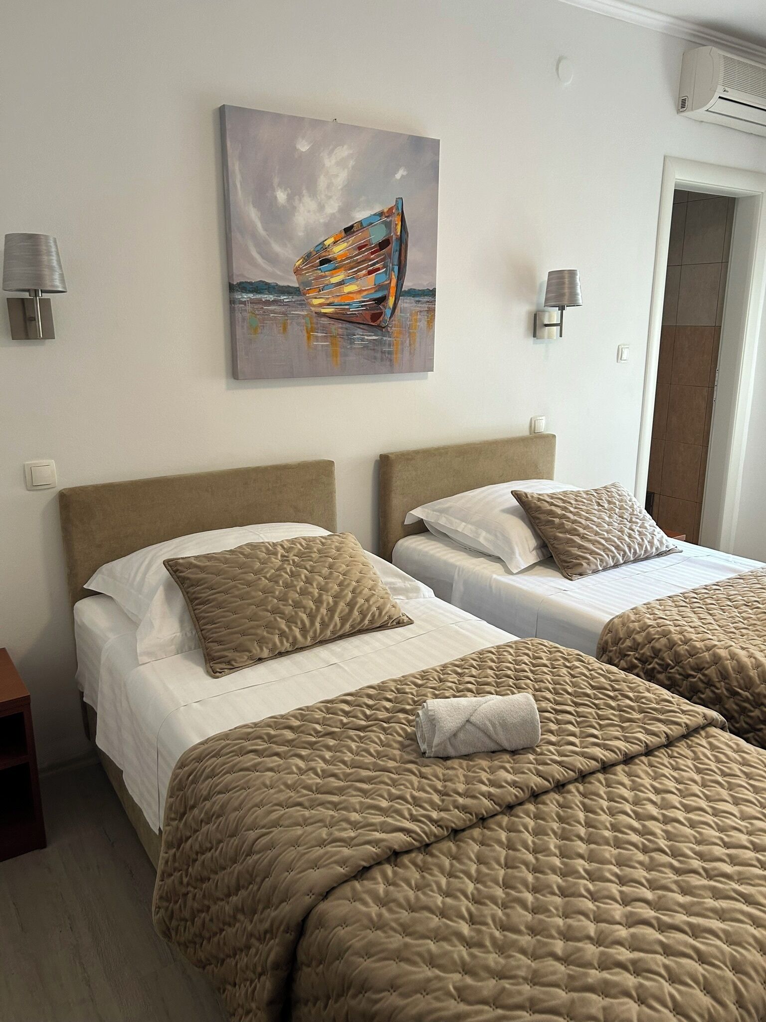 Habitación de hotel con dos camas, ropa de cama color beige, un cuadro de un barco y un aire acondicionado de pared.
