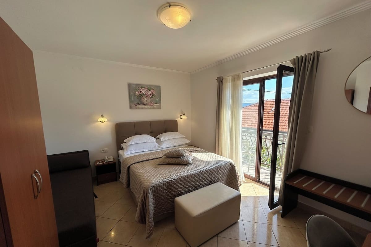 A bedroom with a bed, bedside tables, a desk, a chair, a balcony with a view, and a painting on the wall.