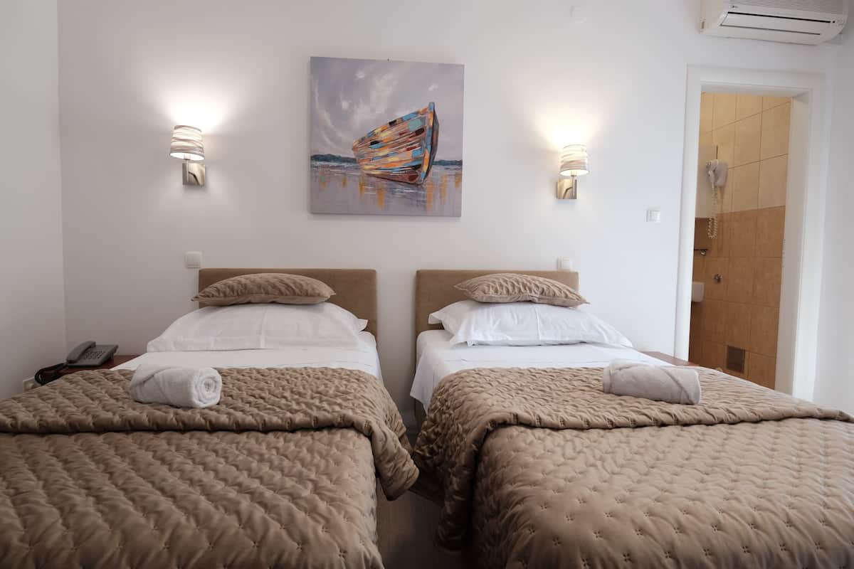 A hotel room with two beds, beige bedding, a painting of a boat, and wall-mounted lights.