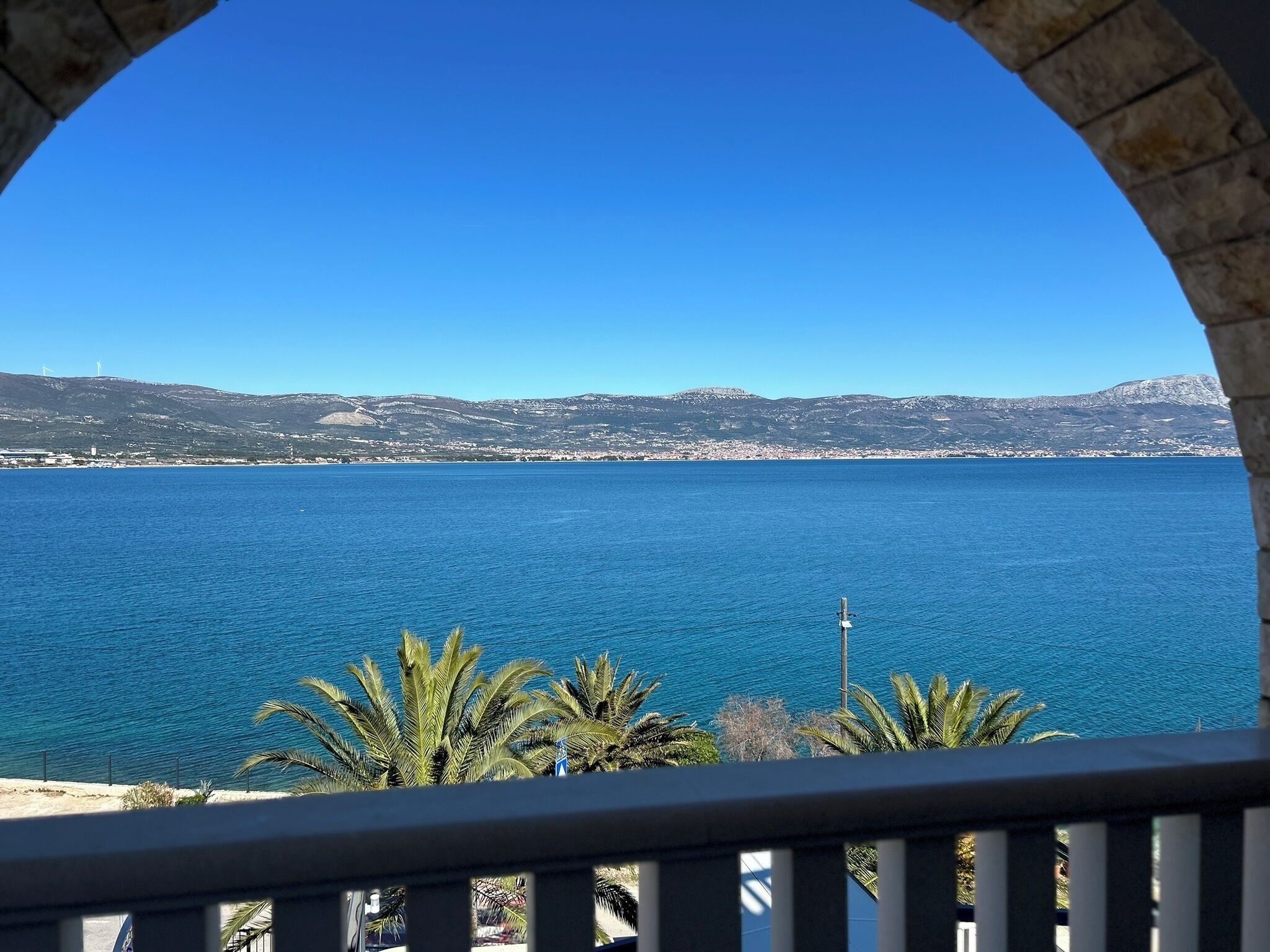 A view through an arched window overlooking a calm sea with distant hills and a clear sky.