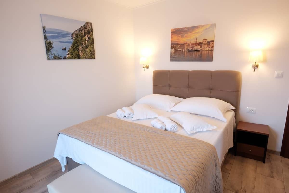 A neatly made bed with rolled towels, a headboard, two wall-mounted lamps, and two framed pictures on the walls.