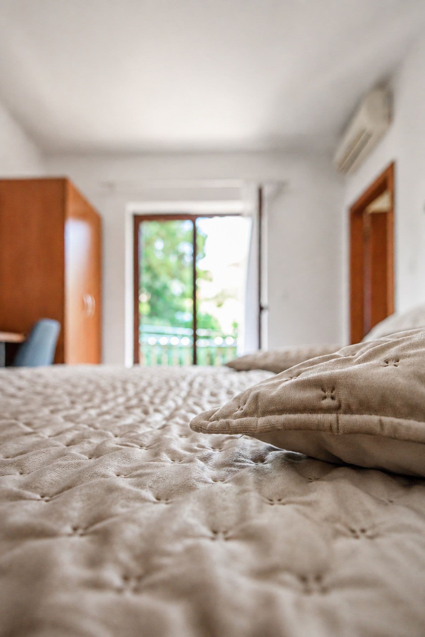 A bed with a textured duvet, a wooden wardrobe, and a view of a balcony with greenery.