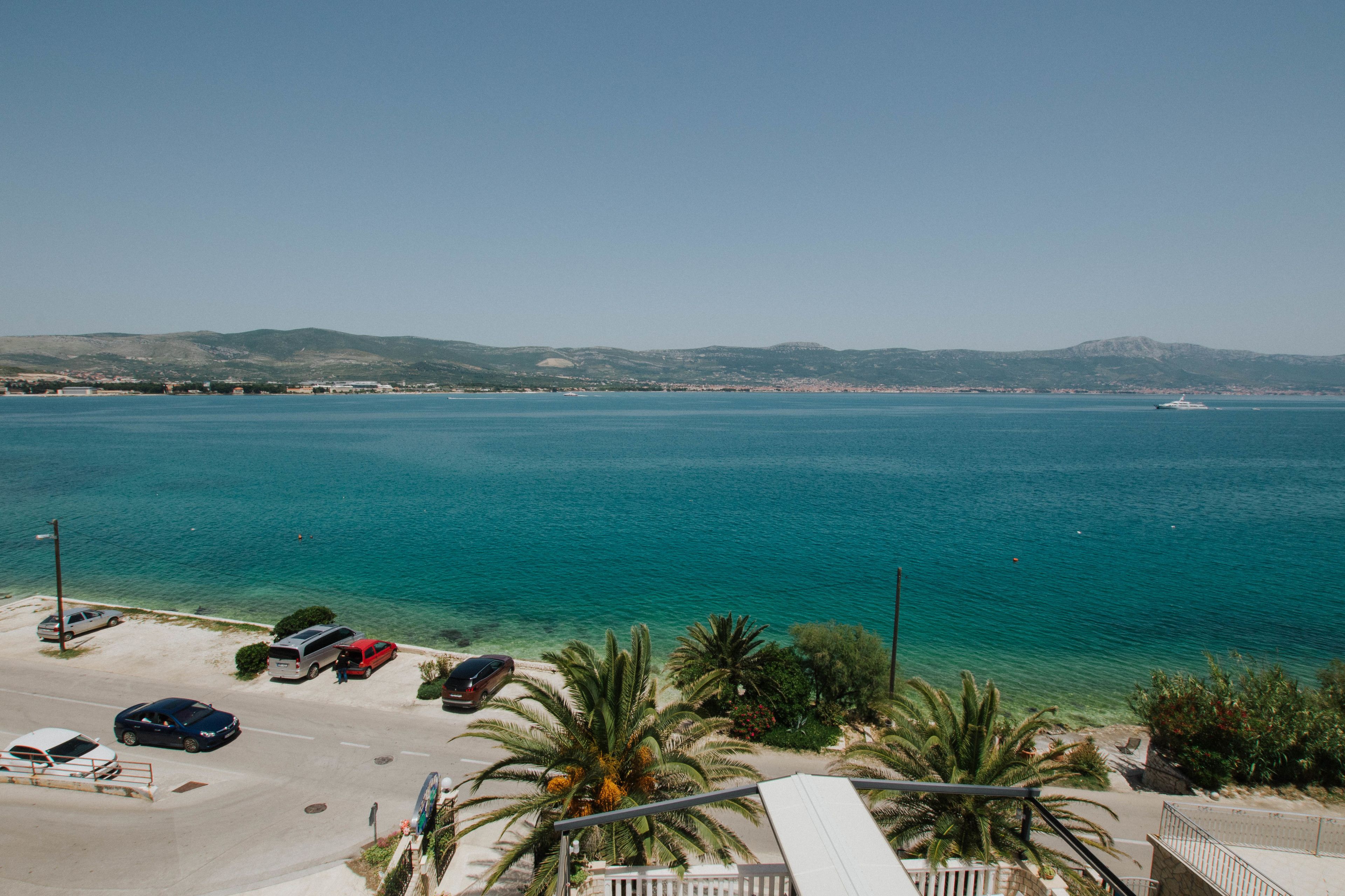 A coastal view with a parking area, vehicles, and a clear blue sea.