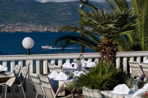 Une terrasse avec des tables dressées pour le dîner, offrant une vue sur un plan d’eau et une ville située au loin.