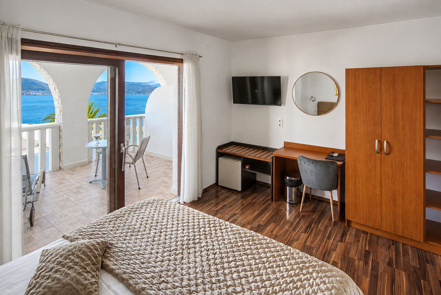 A hotel room with a bed, a desk, a TV, a wardrobe, and a balcony with a view of the sea.