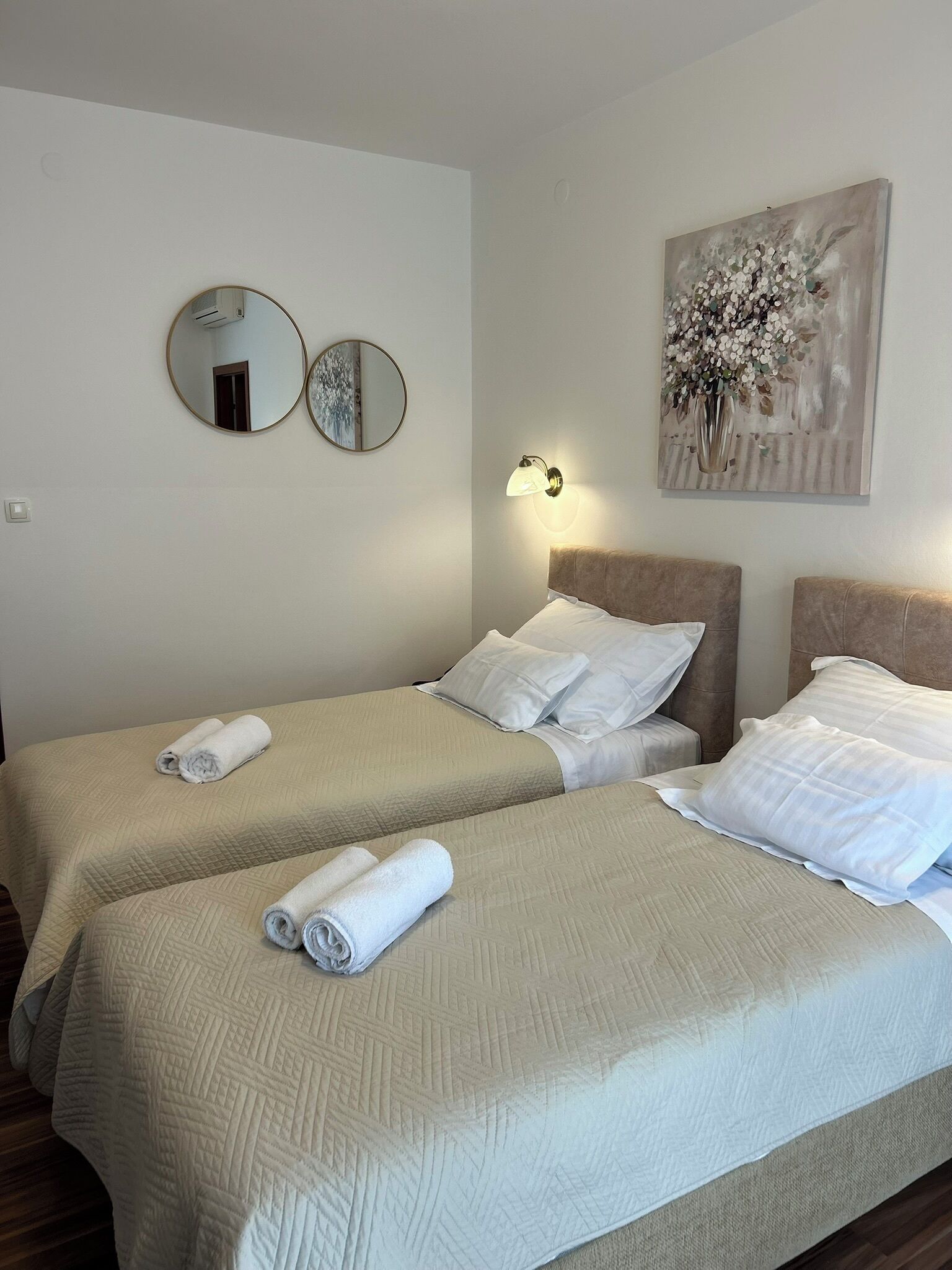 A hotel room with two beds, each with a headboard, a wall-mounted mirror, a wall-mounted light fixture, and a painting of flowers.