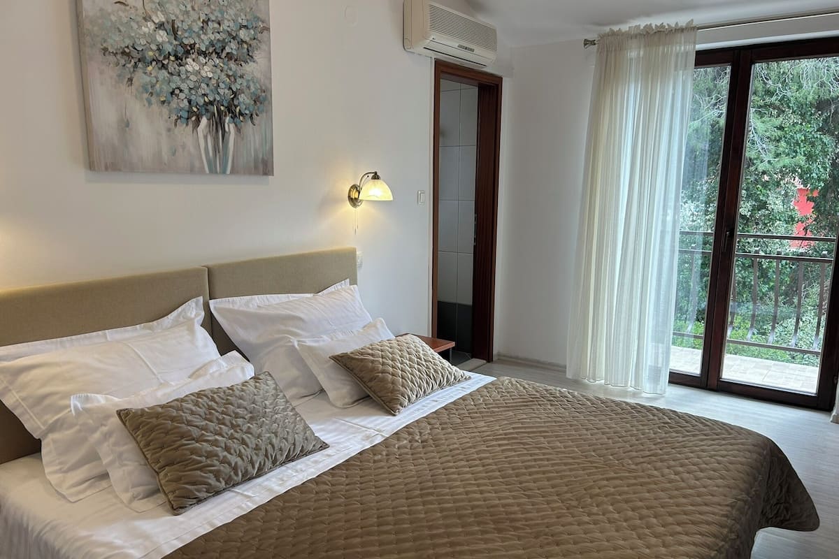 A bedroom with a bed, a painting, an air conditioning unit, and a view through a window.