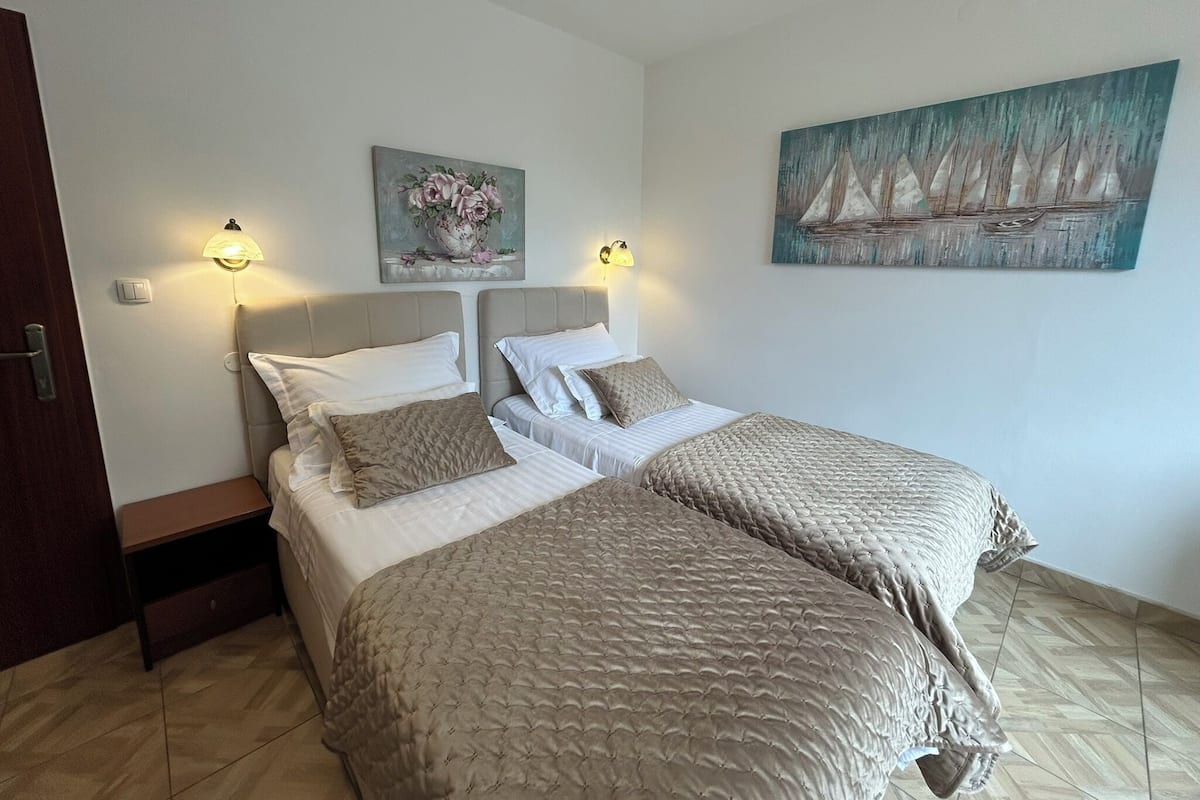 A bedroom with two beds, a nightstand, wall-mounted lamps, and a painting.