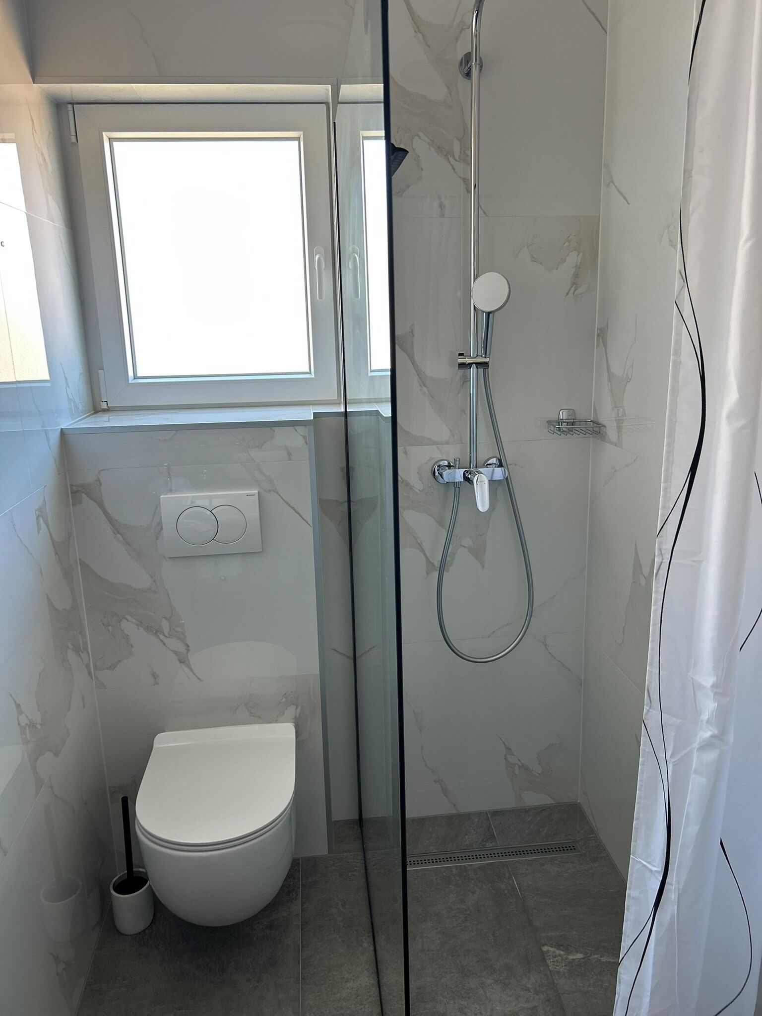 A modern bathroom with a glass-enclosed shower, a toilet, and a window above the sink.