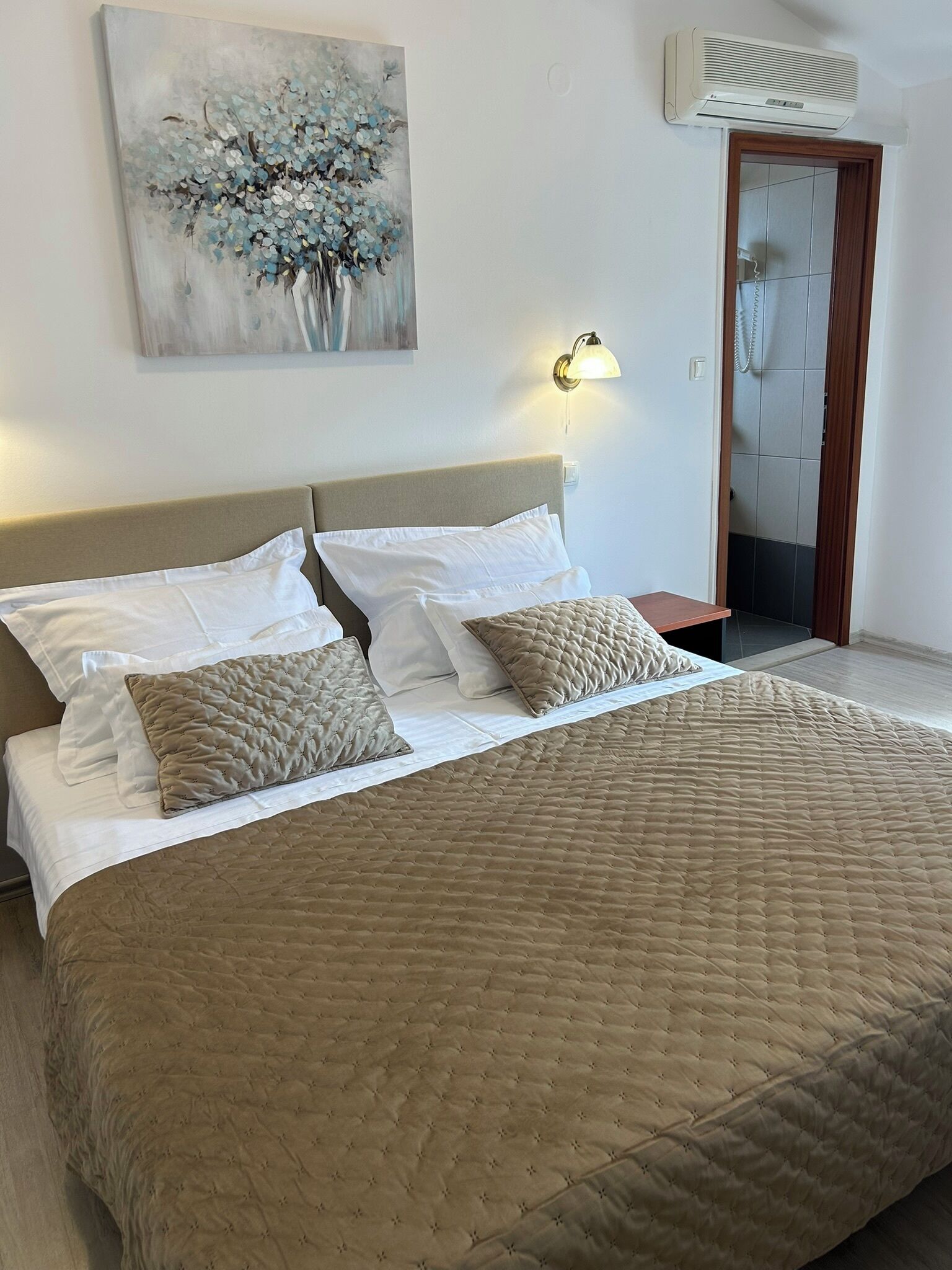 A bedroom with a bed, pillows, a wall-mounted air conditioner, a light fixture, and a door leading to a bathroom.