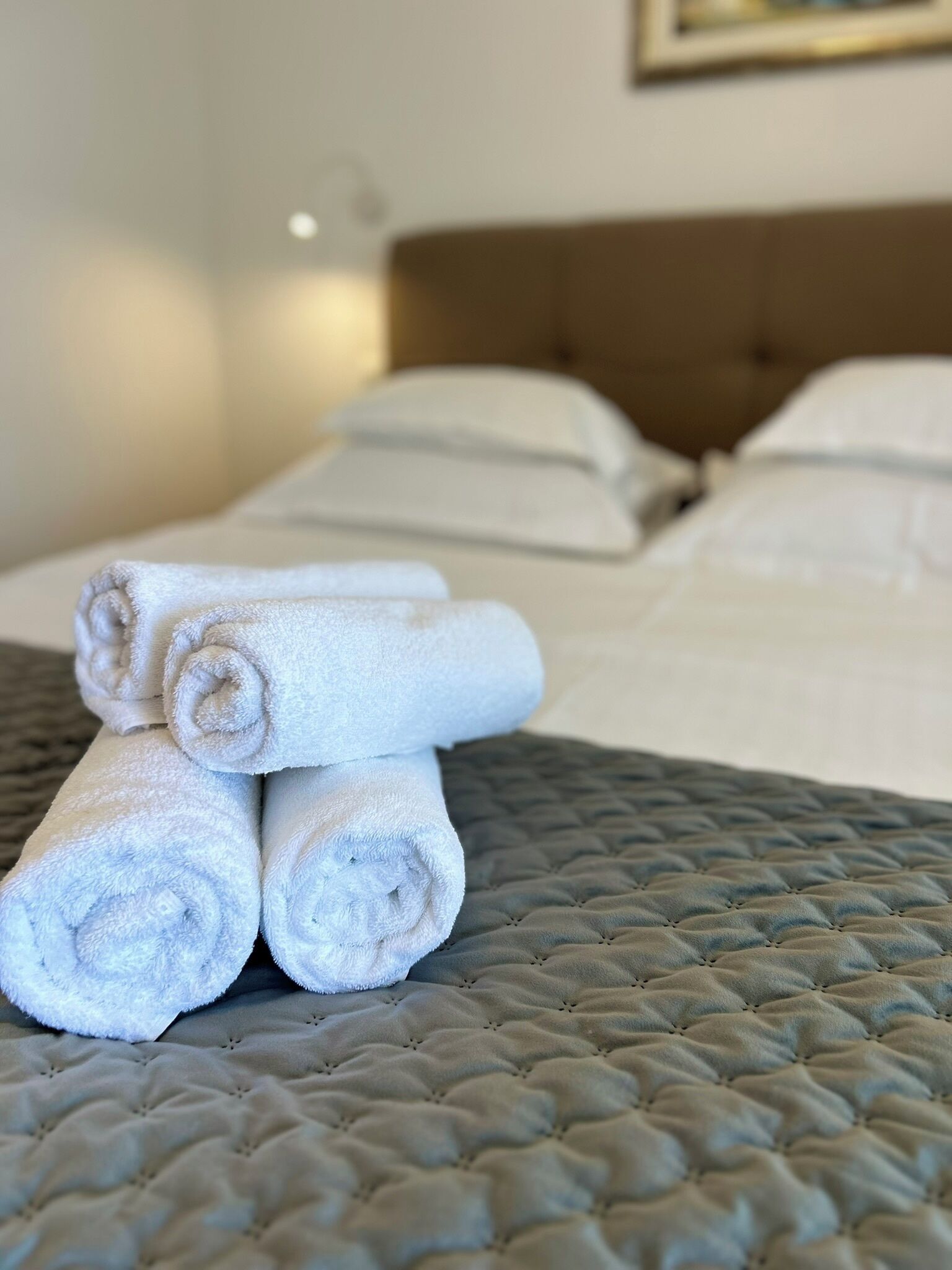 Een net opgemaakt bed met een grijze gewatteerde deken, witte kussens en opgerolde witte handdoeken op het bed.