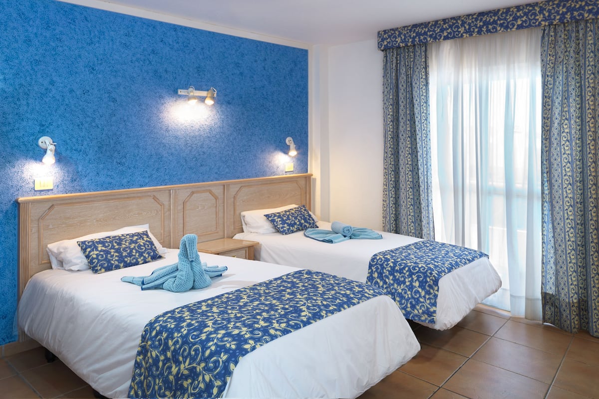 Habitaci ón de hotel con dos camas, una pared azul con textura y un ventanal con cortinas estampadas.