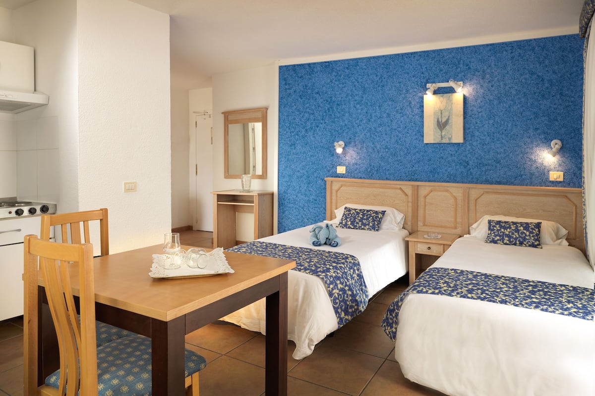 Habitación de hotel con dos camas, una mesa de madera, una silla, una pequeña kitchenette y una pared de acento azul.