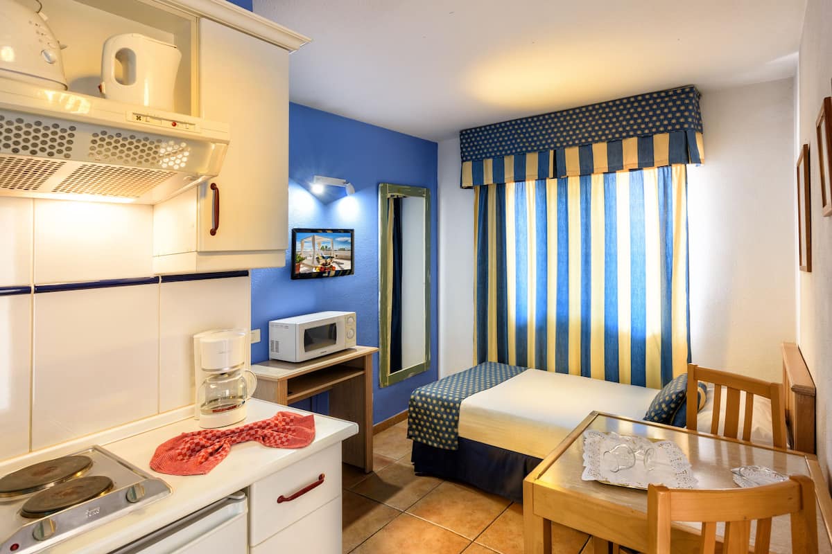 Una habitación de hotel compacta con kitchenette, una cama con cortinas a rayas, una mesita y un espejo.