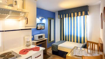 Una habitación de hotel compacta con kitchenette, una cama con cortinas a rayas, una mesita y un espejo.