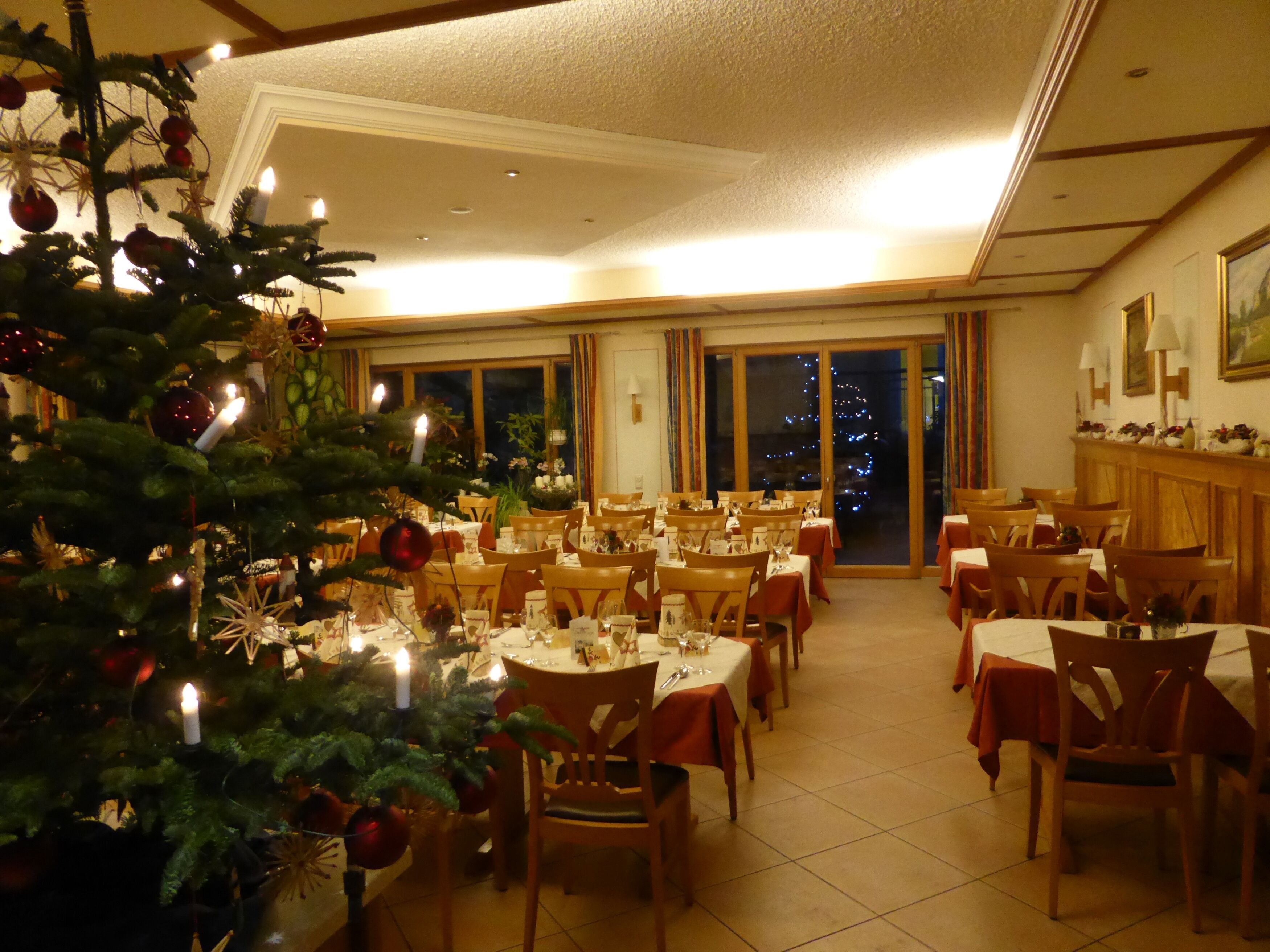 A dining area with tables set for service, a Christmas tree, and a view of the night sky through large windows.