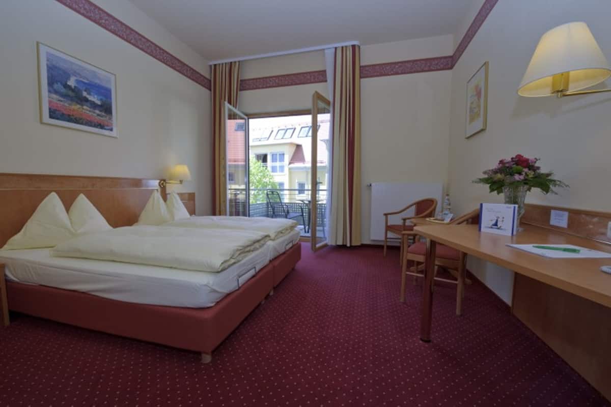 A hotel room with a bed, a desk, a chair, a lamp, and a window with a view of buildings.