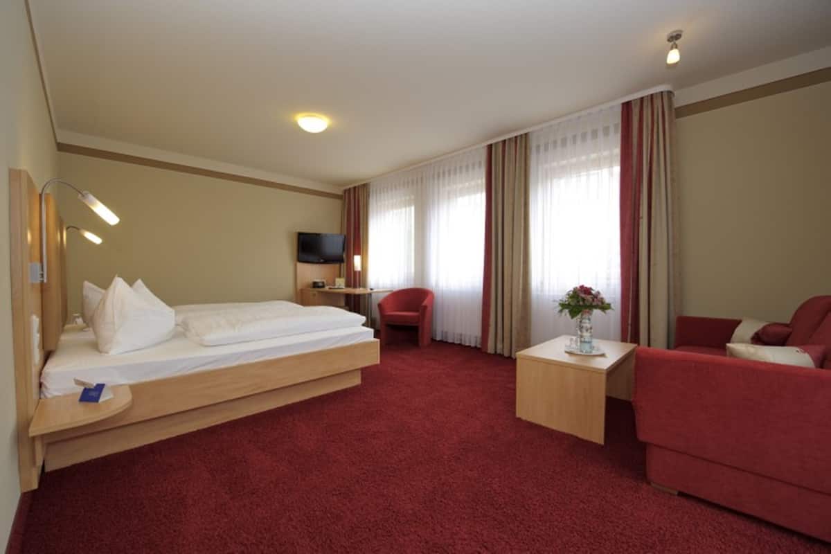 A hotel room with a bed, a desk with a TV, a red armchair, a red sofa, and a small table with a vase of flowers.
