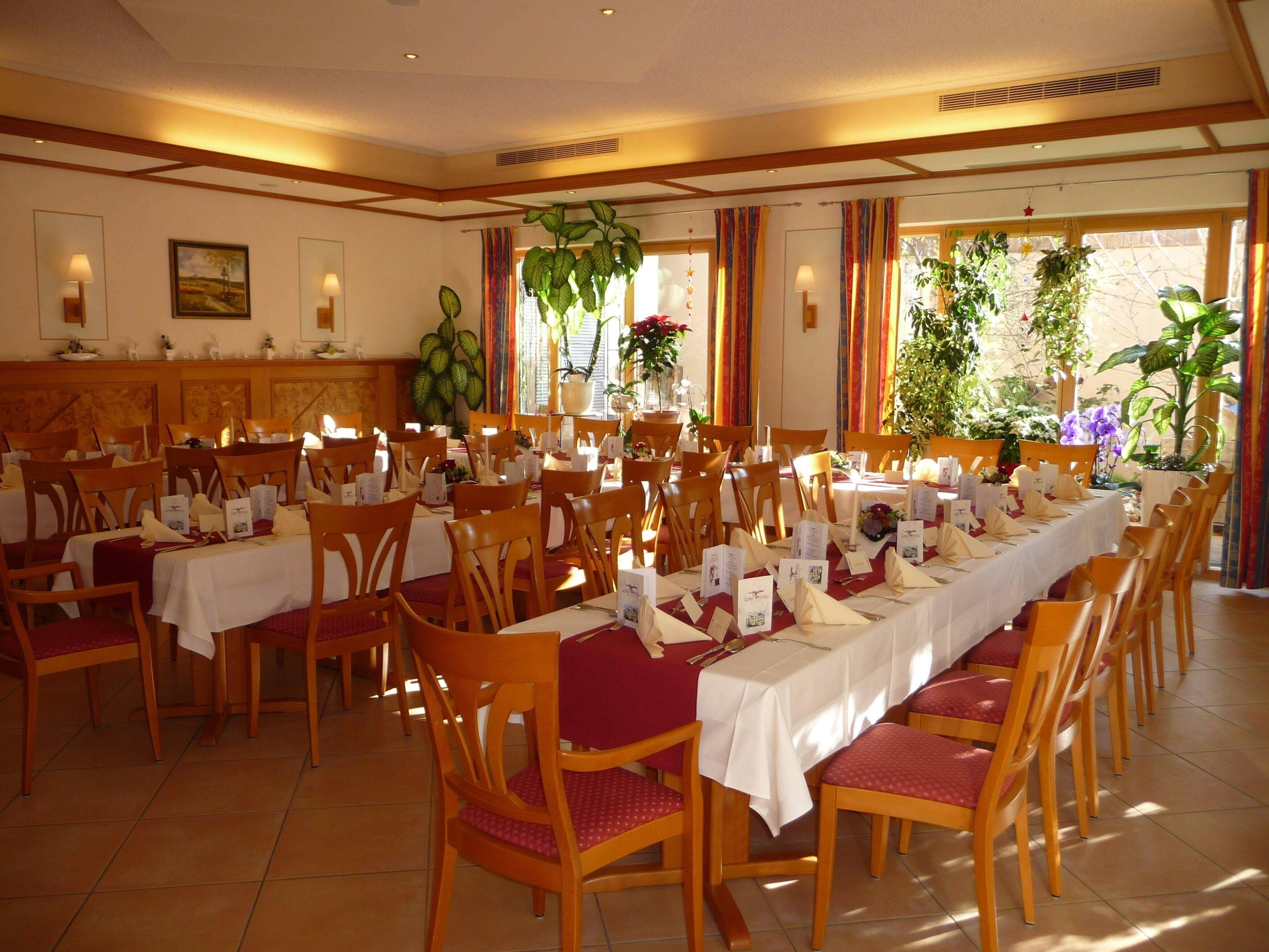 Un comedor amplio con varias mesas y sillas, ventanas grandes con cortinas y plantas en macetas.