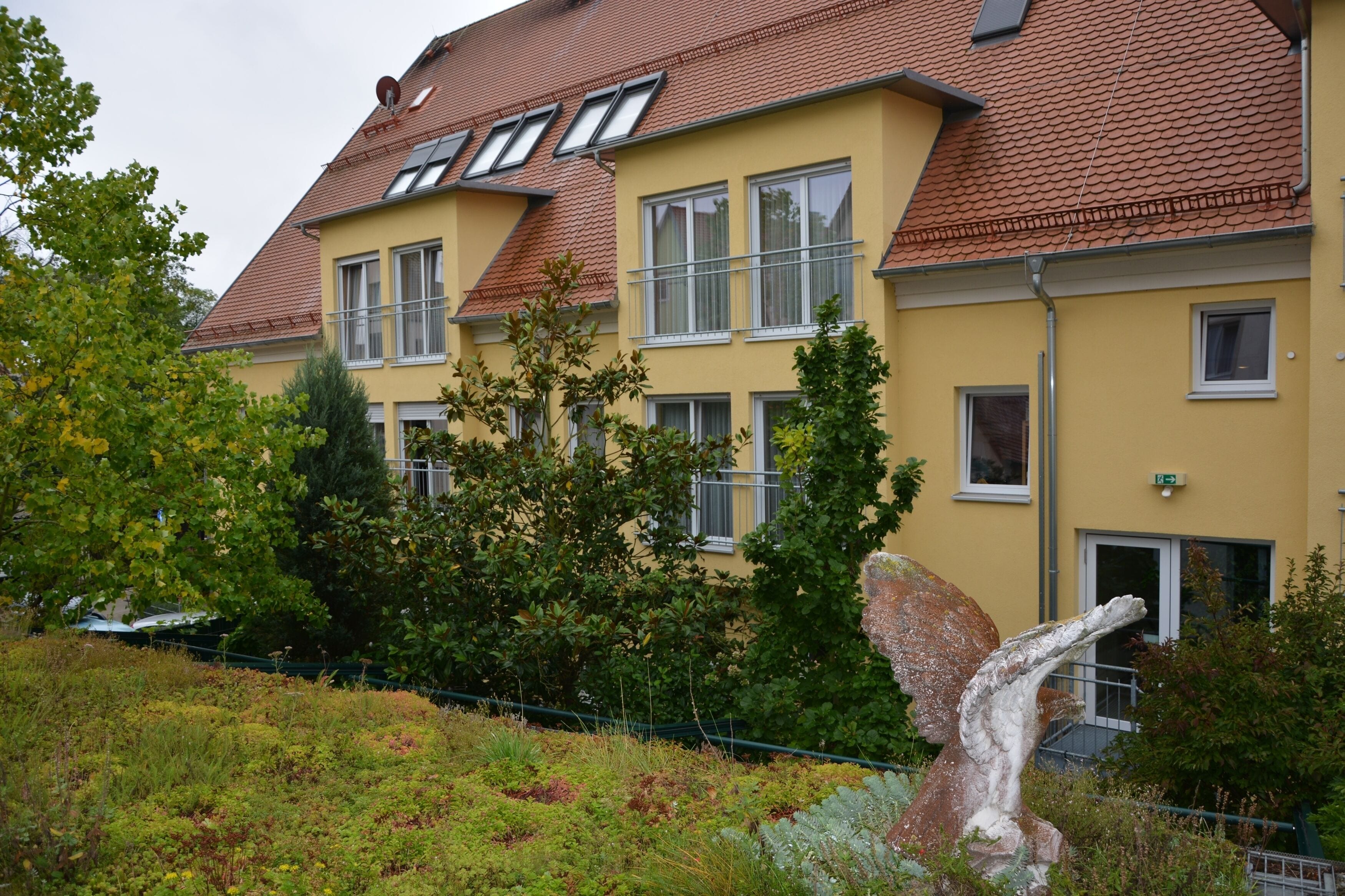 Ein gelbes Gebäude mit vielen Fenstern und einem roten Dach, umgeben von Grünpflanzen und einer Felsenformation.