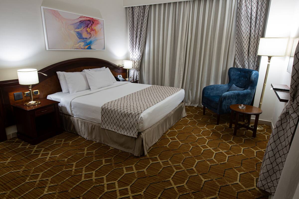 A hotel room with a large bed, a blue armchair, a small table, and a painting on the wall.