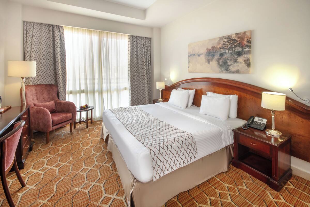 A hotel room with a large bed, a desk, a chair, and a nightstand with a lamp.