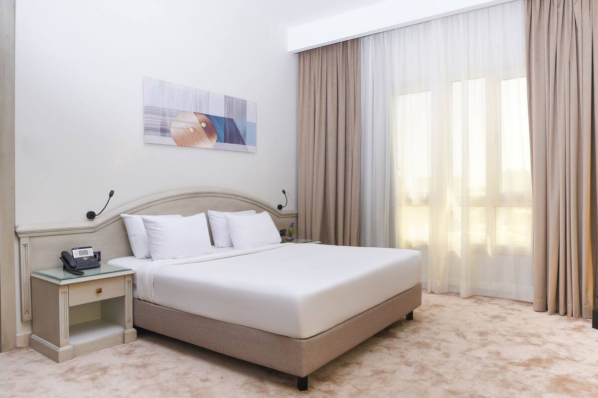 A neatly made bed with a white comforter and pillows, a bedside table with a telephone, and a painting on the wall.