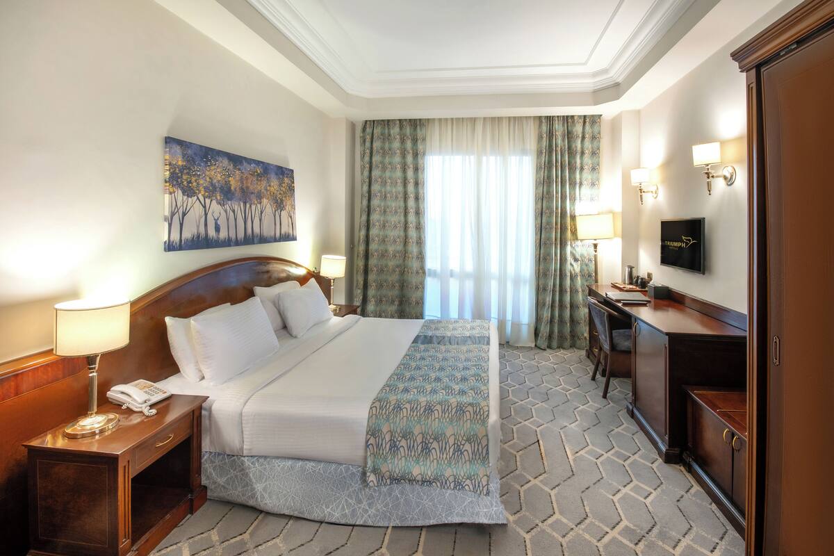 A hotel room with a large bed, bedside table with a lamp, a desk with a chair, a television, and a painting on the wall.