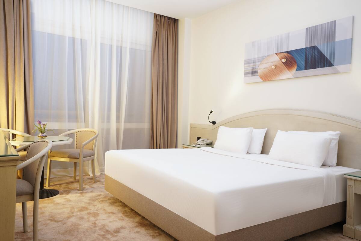 A hotel room with a large bed, a desk with a chair, a telephone, and a painting on the wall.