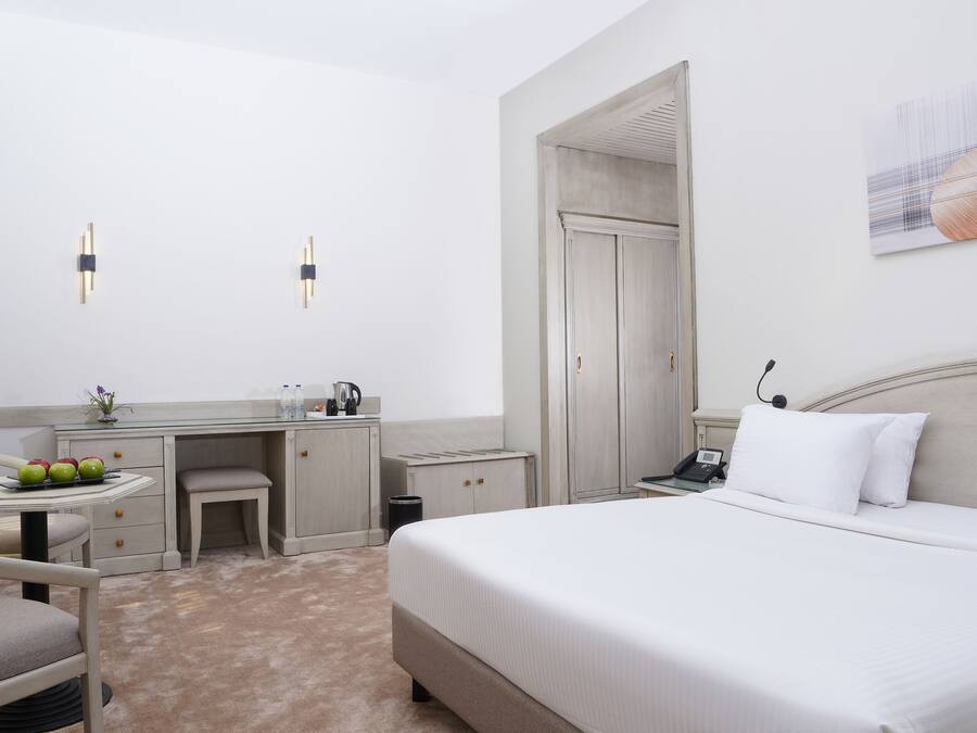 A hotel room with a large bed, a desk with a chair, a small table with apples, and a trash bin.
