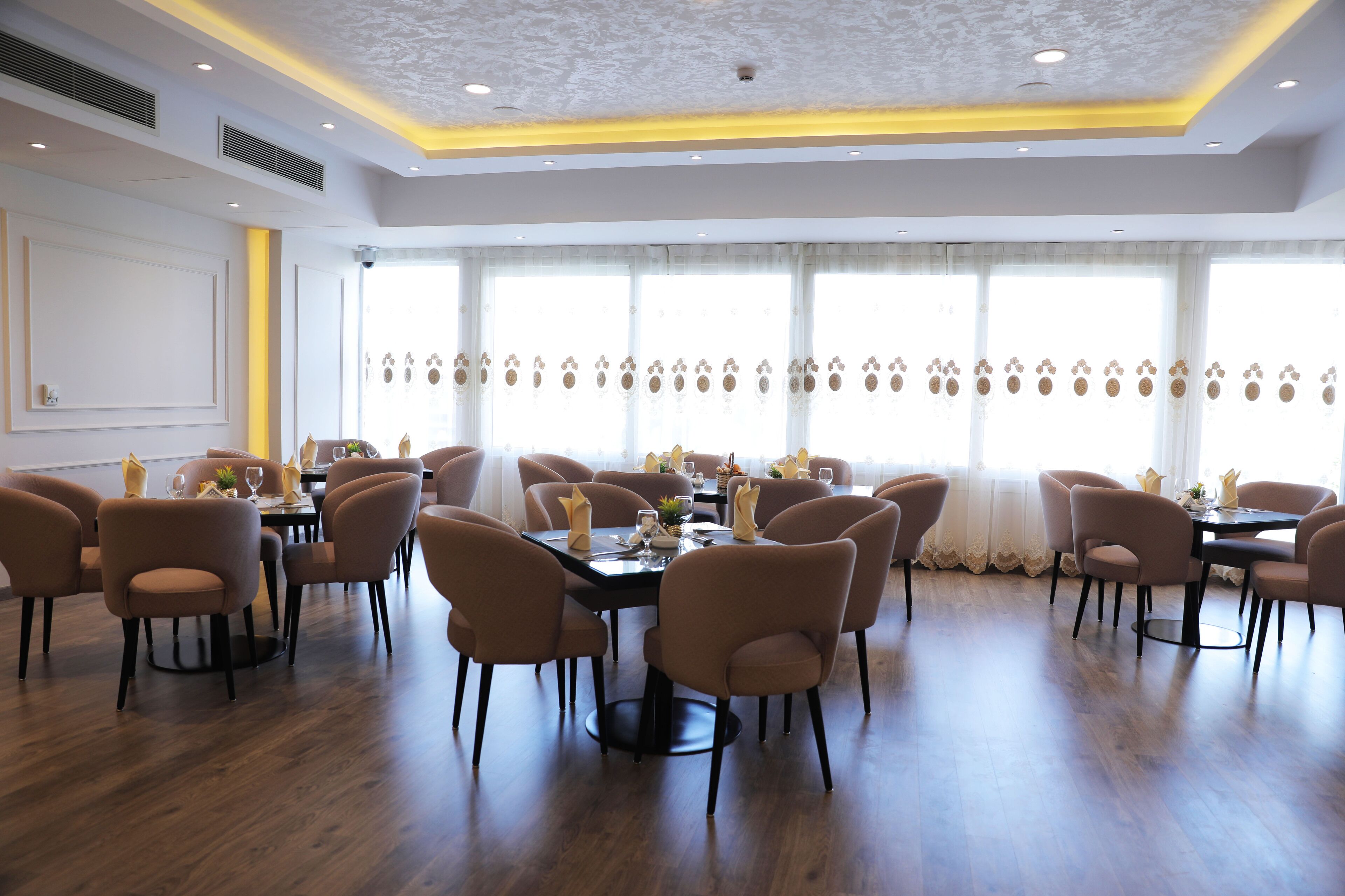 A spacious dining area with multiple tables and chairs, large windows with curtains, and a modern ceiling design.