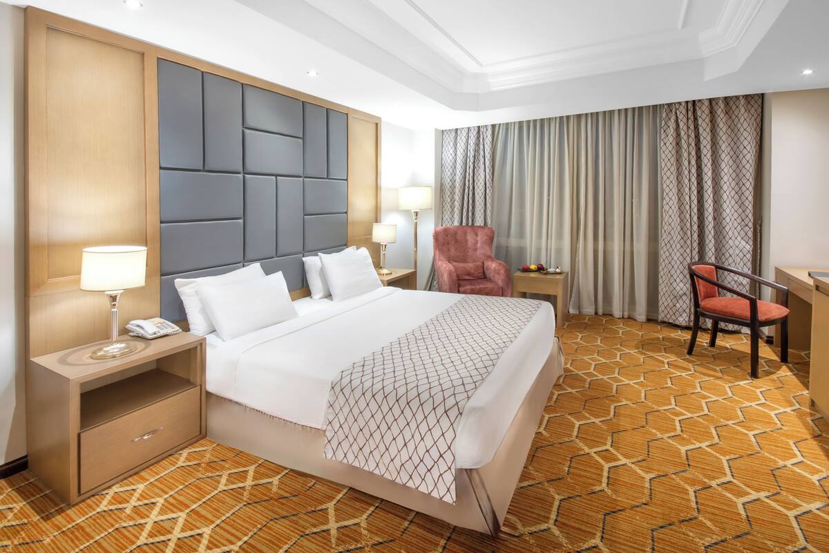 A hotel room with a large bed, a desk, a chair, and a nightstand with a lamp.