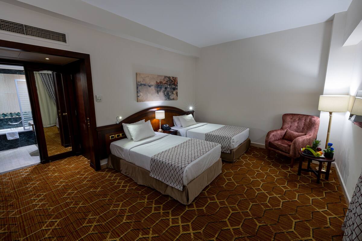 A hotel room with two beds, a sitting area with a chair, a small table with a lamp, and a large window.