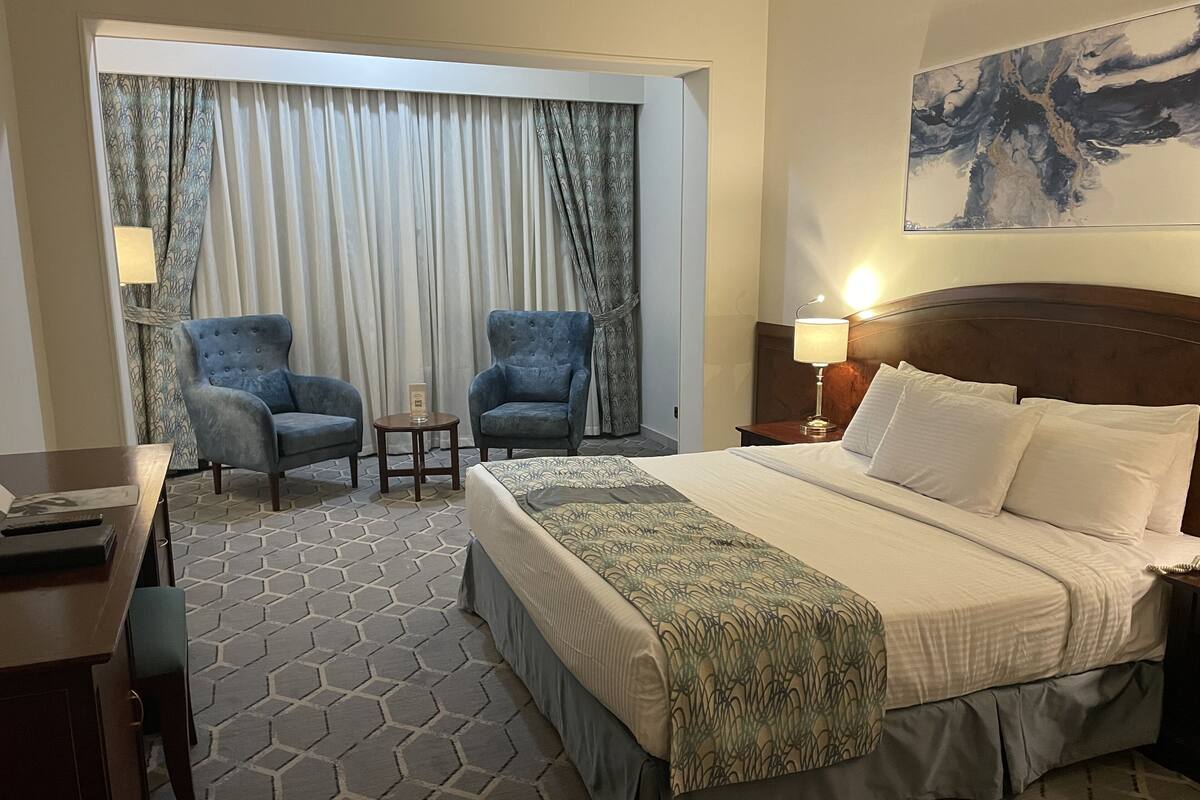 A hotel room with a large bed, two armchairs, a small table, and a nightstand with a lamp.