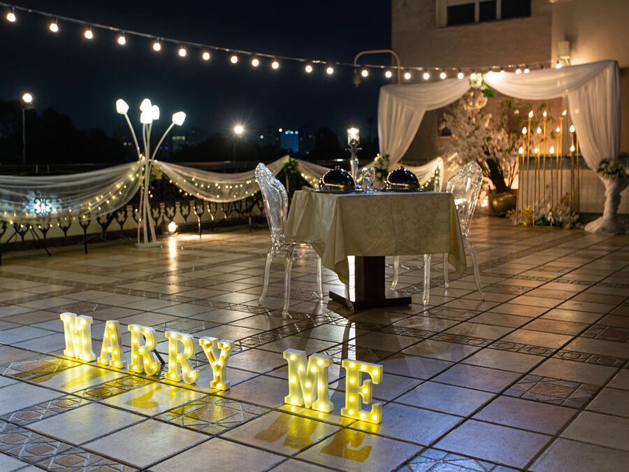 Un cadre romantique en plein air, avec une table dressée pour deux, recouverte d’un tissu blanc et illuminée par des guirlandes lumineuses.