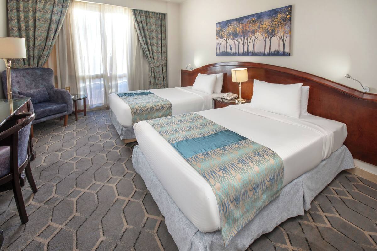 A hotel room with two beds, a desk, a chair, and a painting on the wall.