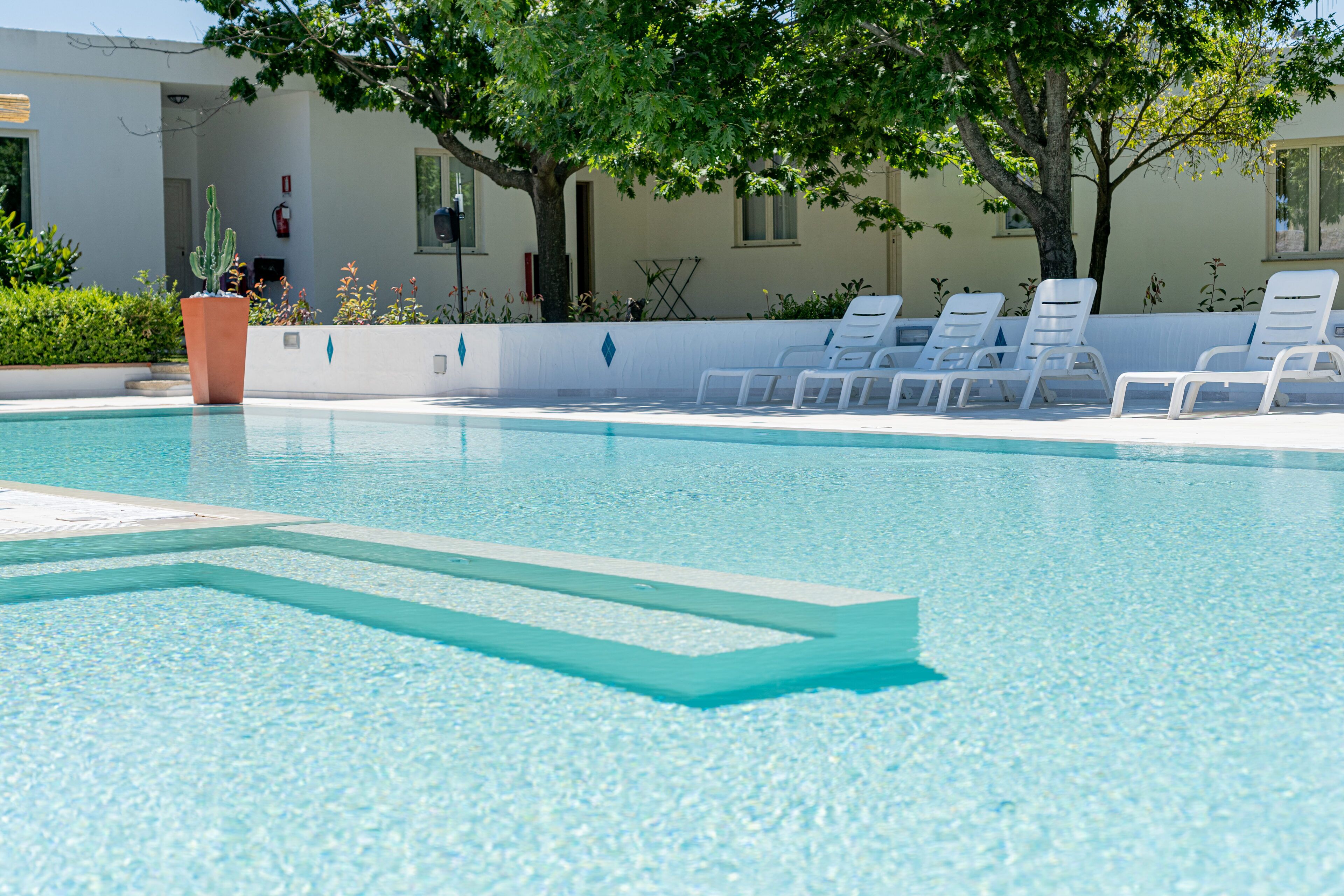 A clear swimming pool with a tiled deck, surrounded by white lounge chairs and a potted cactus.
