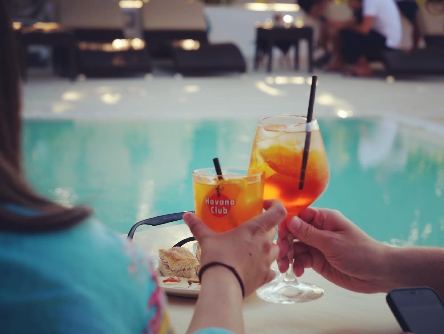 Zwei Personen stoßen mit Getränken an einem Tisch am Pool mit Blick auf das Wasser auf.