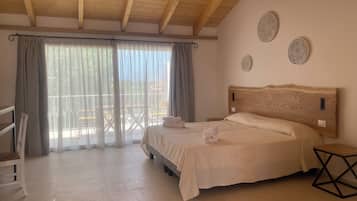 Une chambre avec un plafond en bois, un lit recouvert de draps blancs, une petite table de chevet et une vue dégagée sur l’extérieur grâce à une grande fenêtre ornée de rideaux légers.