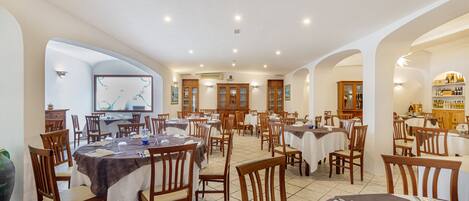 A spacious dining area with multiple tables and chairs, wooden cabinets, and a large screen displaying a map.