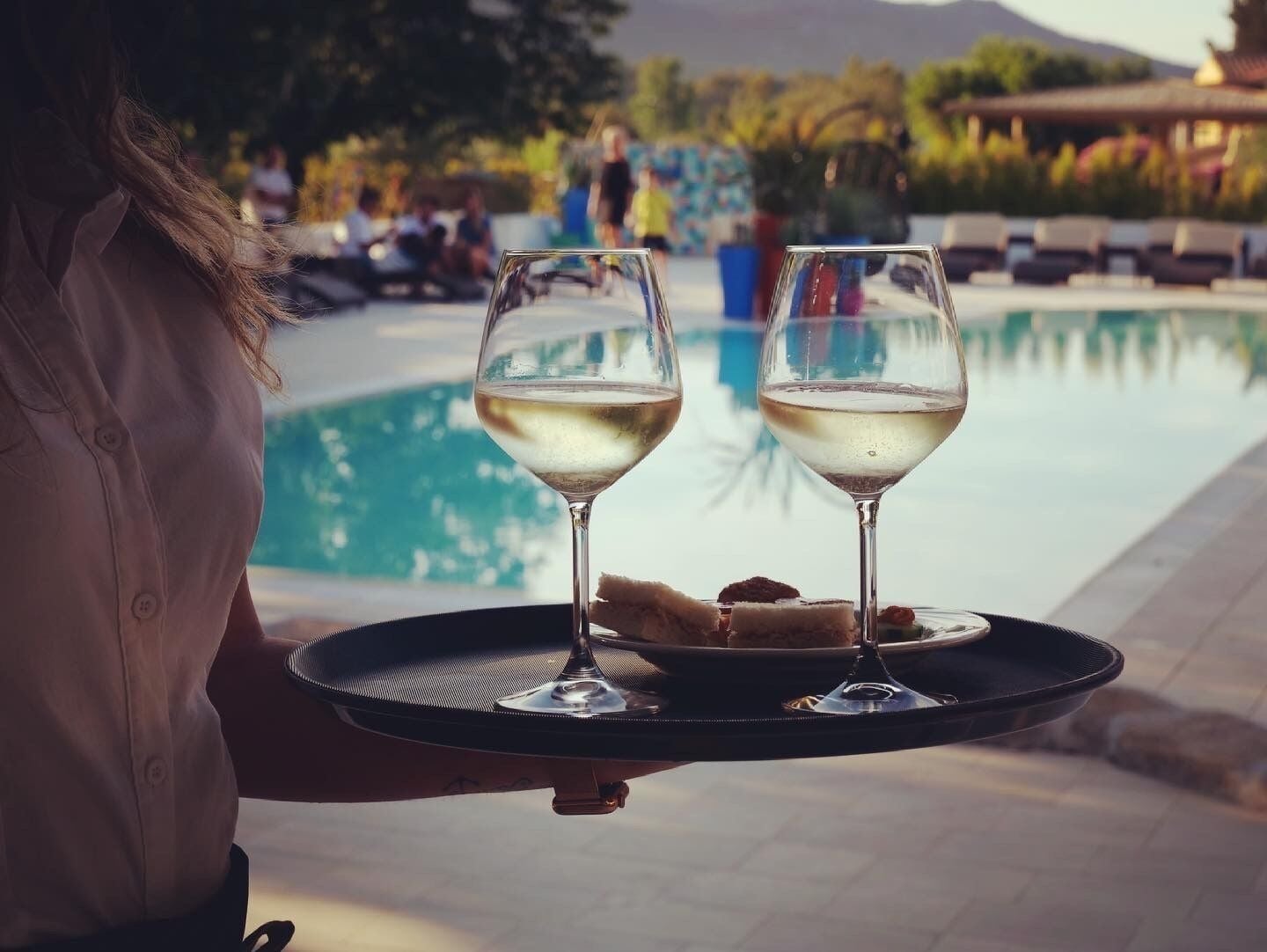 Une personne tenant un plateau avec deux verres de vin blanc et une assiette de nourriture, près d’une piscine.
