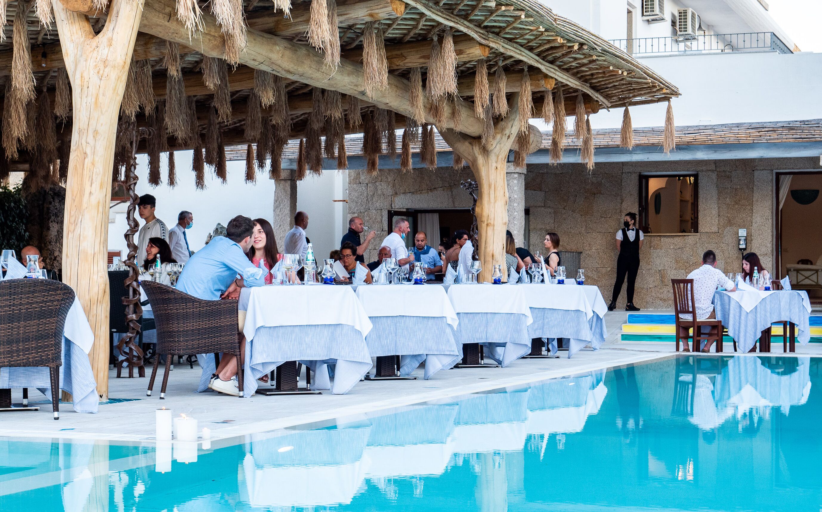 An outdoor dining area with tables set up by a poolside, featuring a wooden pergola overhead.