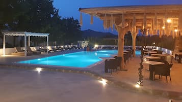 A poolside area with lounge chairs and tables, a covered seating area, and a view of mountains in the background.