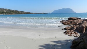 Ein Strand mit klarem, blauem Wasser, felsiger Küste und einem entfernten Berg.