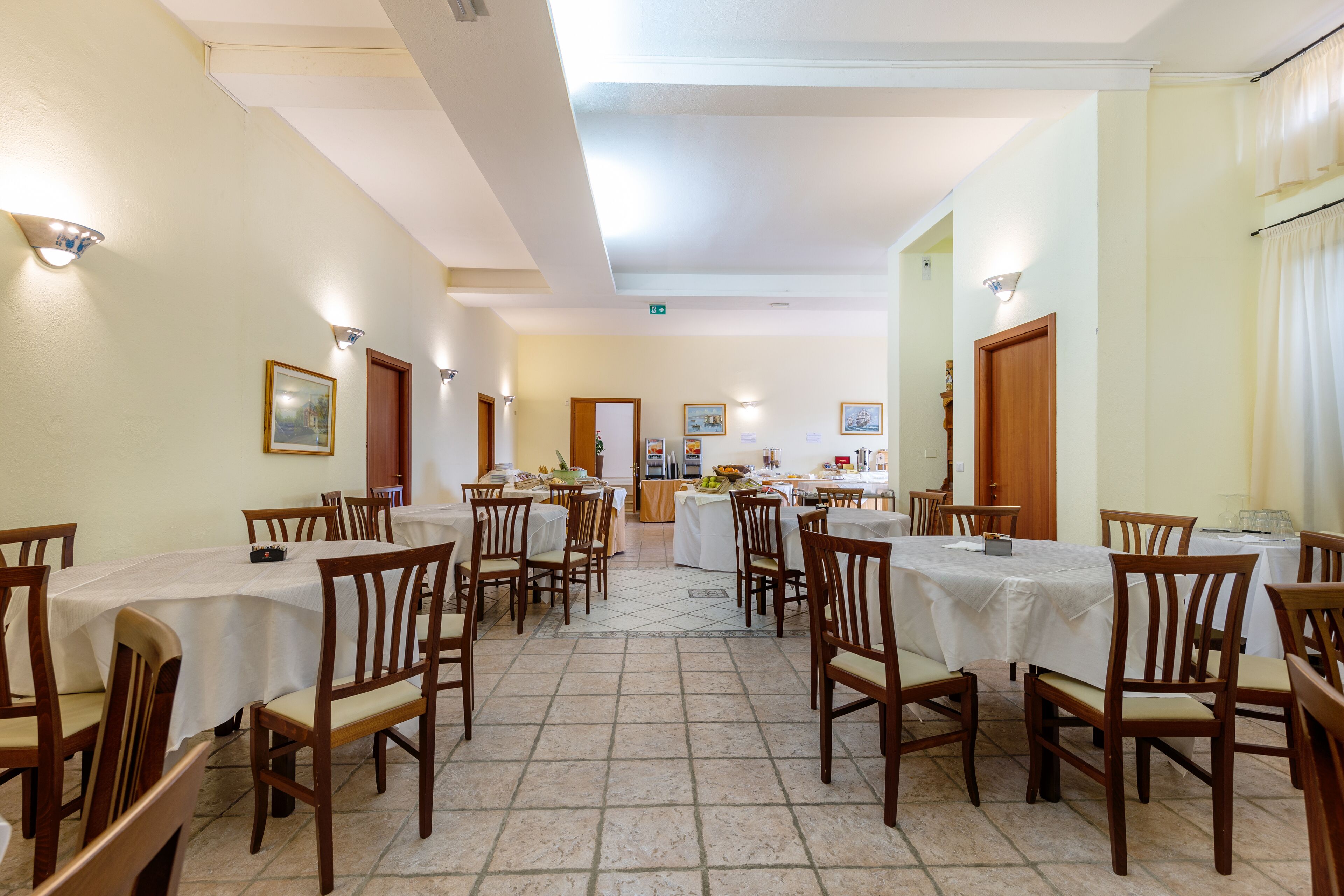Un comedor con varias mesas y sillas, suelo de baldosas y cuadros en las paredes.